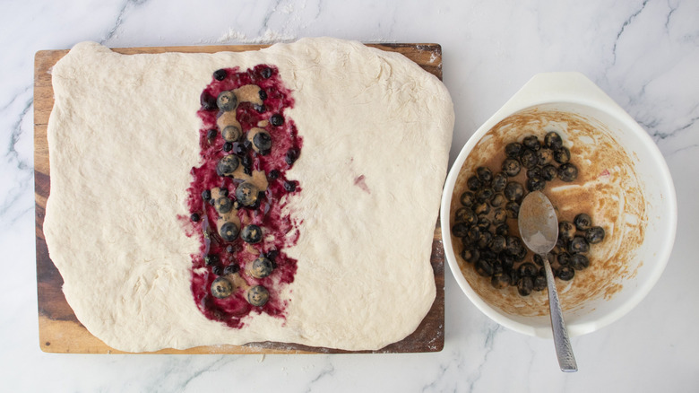 Dough rectangle topped with blueberry jam and spiced mixture next to bowl of blueberry mixture