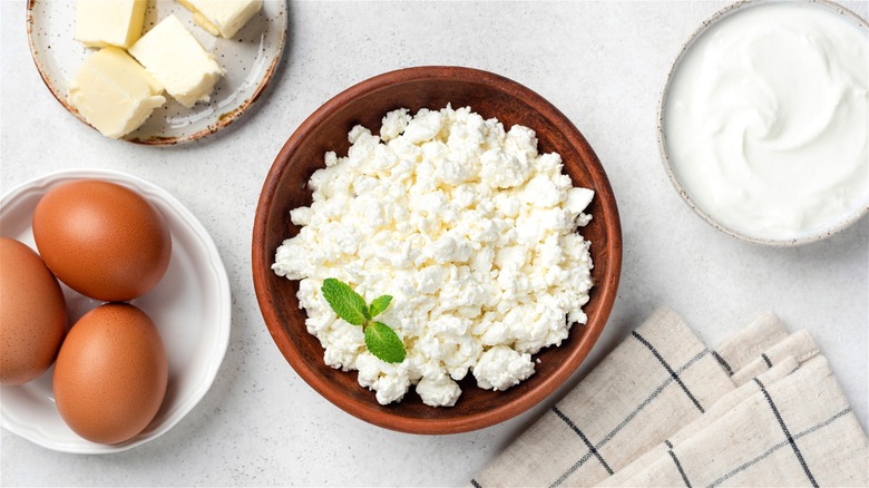 Cottage cheese in a wooden bowl, with eggs, butter, yogurt, and napkins displayed around it.
