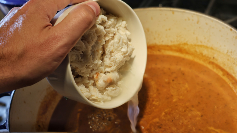 hand dumping bowl of lump crab meat into pot of red soup