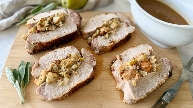 Pear-stuffed pork loin pieces on wooden board with gravy boat and sage