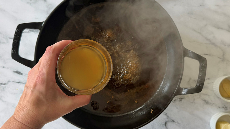 Hand holding bowl of chicken stock over steaming skillet