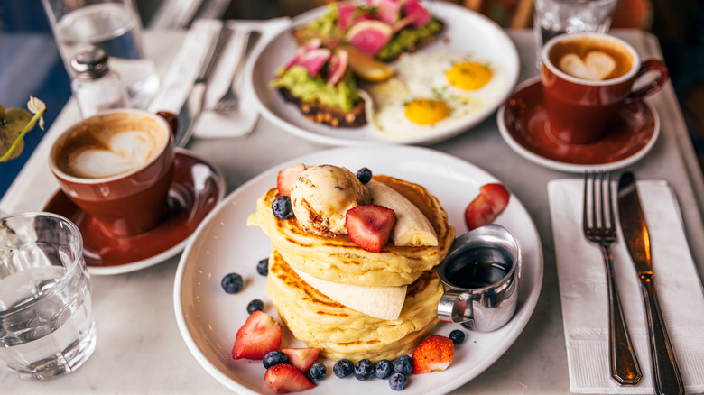 stack of pancakes with fruit, sunny side up eggs with avocado toast, and two coffees