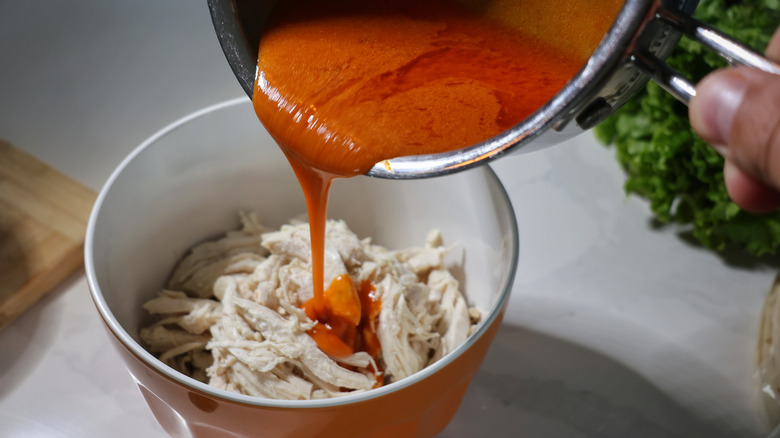 Buffalo sauce pouring from saucepan into mixing bowl of shredded chicken