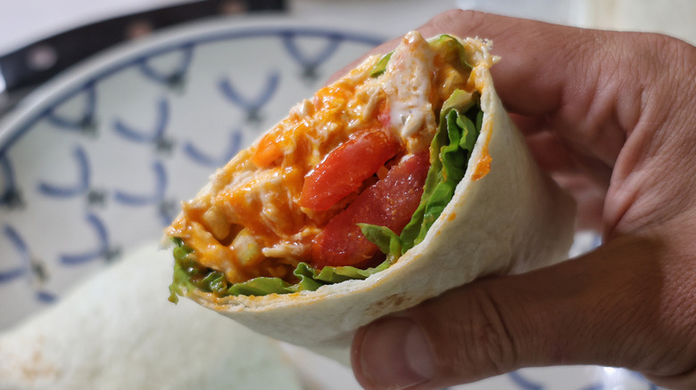 hand holding half of a shredded Buffalo chicken wrap with lettuce and tomatoes