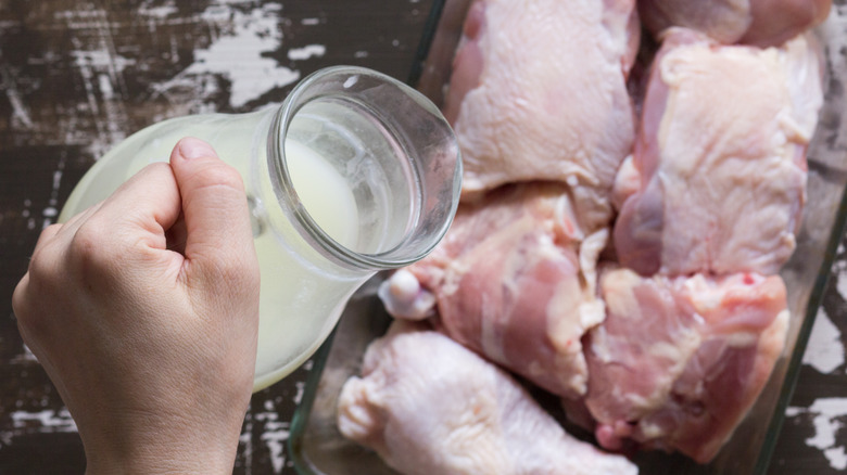 Hand pouring jug of milk onto raw meat.