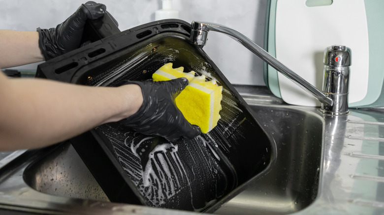 Person wearing gloves washing air fryer basket in sink.