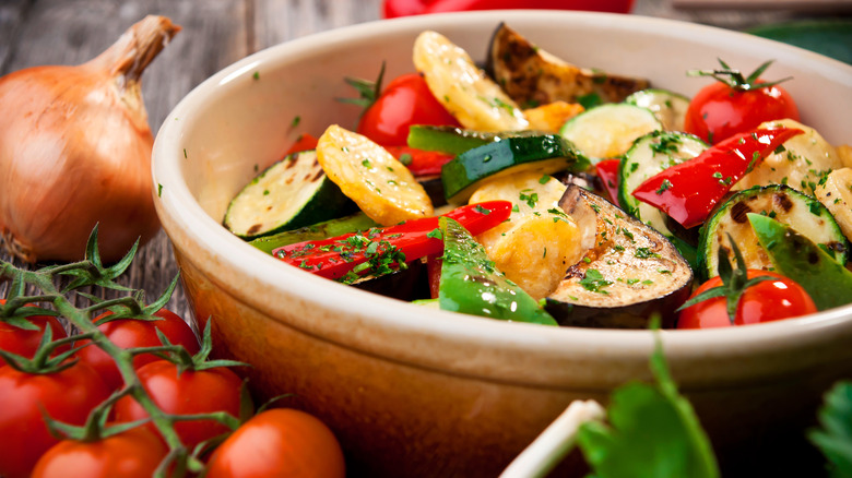 colorful bowl of cooked vegetables including zucchini and red peppers with fresh herbs on wooden table with raw onion and tomatoes alongside