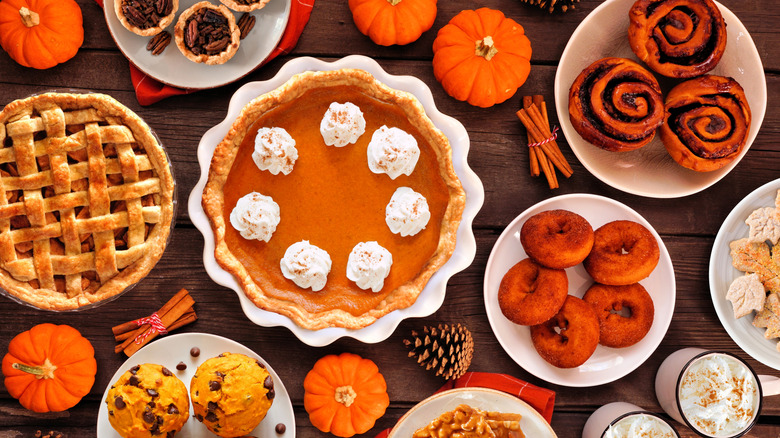 A variety of autumn desserts such as pumpkin pie, apple pie, cinnamon buns, and donuts on a wooden table.