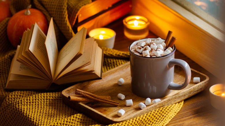 A mug of hot chocolate on a tray surrounded by candles and an open book.