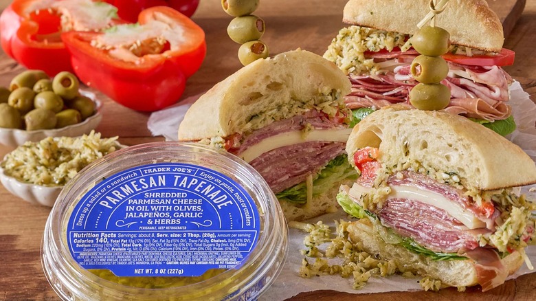Sliced deli sandwiches sit next to a container of parmesan tapenade with ingredients in the background.