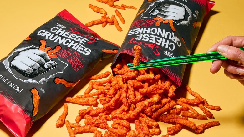 Person eating cheese snacks with chopsticks with two bags of snacks spread out on table.