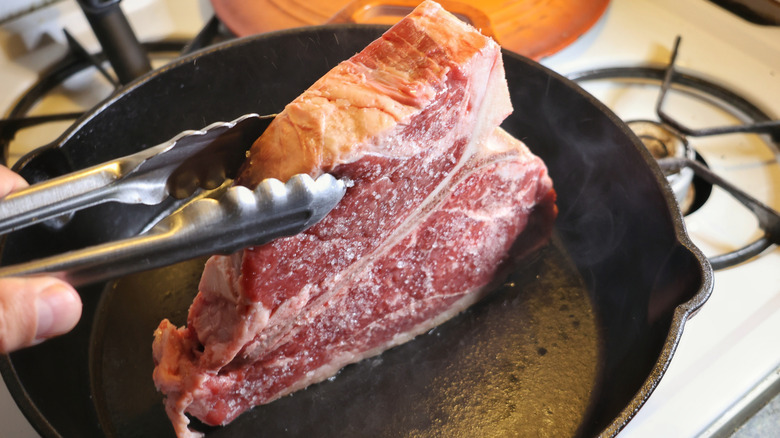 tongs holding t-bone steak on its side in cast iron pan