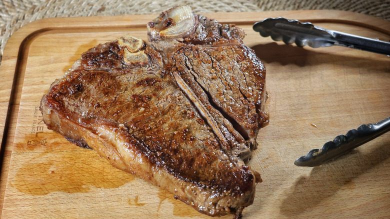 cooked t-bone steak resting on wooden cutting board next to metal tongs