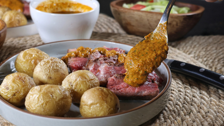 spoon drizzling chimichurri rojo onto thinly-sliced rare steak on plate with roasted potatoes