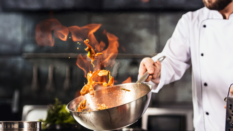 Chef in white coat holding a wok with flame