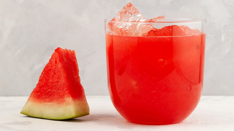 Watermelon cocktail in glass next to watermelon slice on white table with gray background.