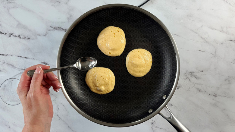Hand holding spoon over pan with pancakes cooking