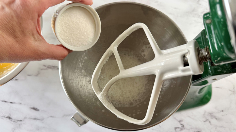 Small bowl of sugar going into stand mixer bowl with paddle attachment