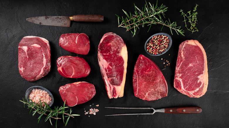Various cuts of beef with fork, knife, and herbs on stone background
