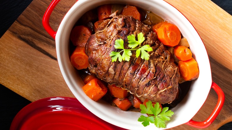 A Dutch oven containing a pot roast and carrots, served on a wooden board.