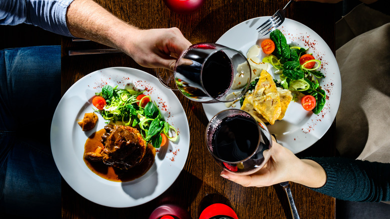 two people clinking red wine glasses over plates of salad and meat