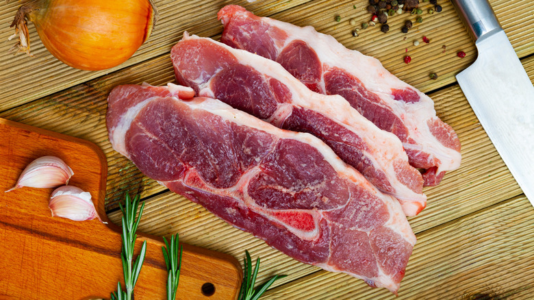 Raw pork shoulder fillets on a cutting board, surrounded by an onion, whole garlic, and herbs.