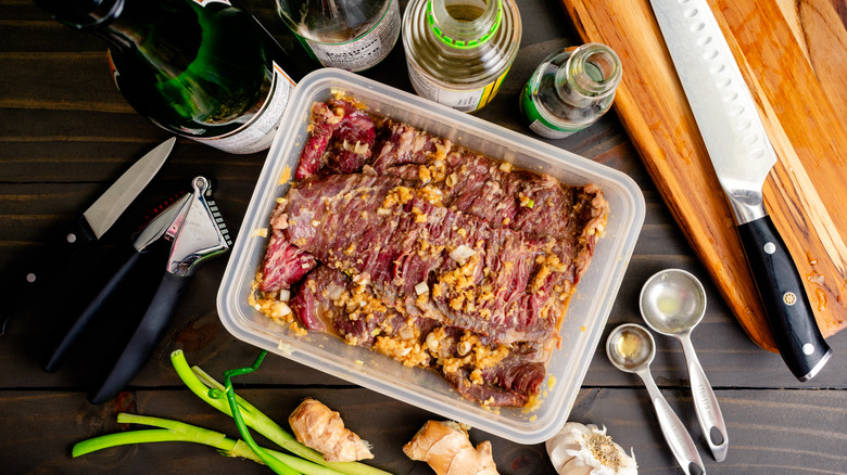 Marinating steak in plastic container with glass bottles of condiments along with herbs and utensils