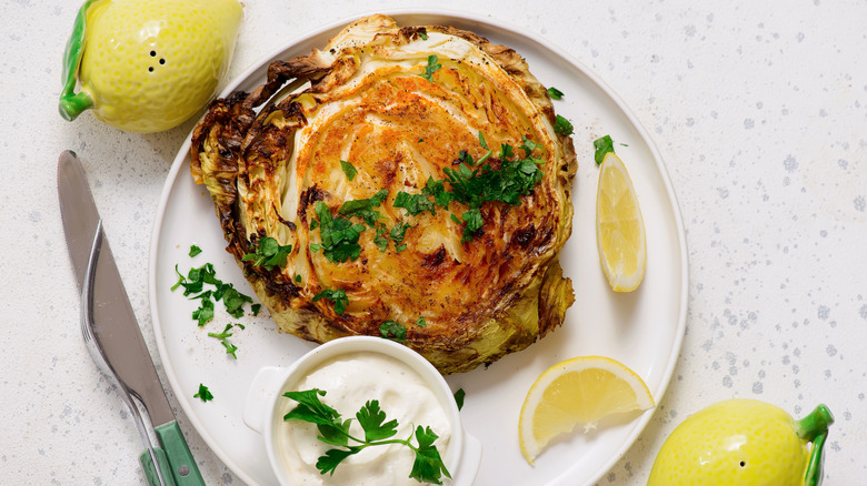 Roasted cabbage with chopped parsley on white plate with lemon wedges and sour cream in white bowl.