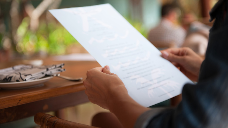 Guest reading paper menu at a restaurant with a wooden table and a white plate with cloth napkin