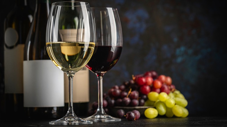Glasses of red and white wine on a table, with their respective bottles and grapes in the background.