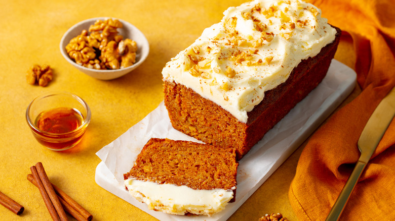 A loaf of carrot cake with walnuts, honey, and cinnamon sticks next to it