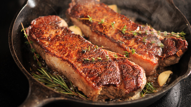 two New York strip steaks being seared in a cast iron pan with herbs and butter