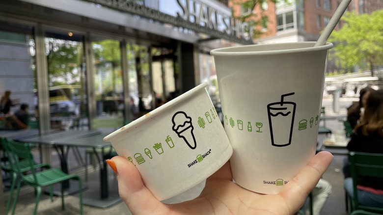 A hand holding a cup of shake shack frozen custard and a shake in front of a shake shack restaurant location