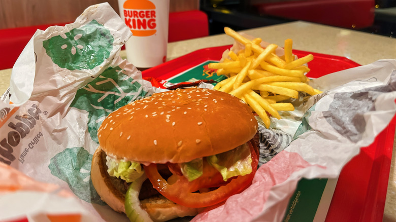 A Buger King Whopper in a paper wrapper next to french fries and a soft drink cup