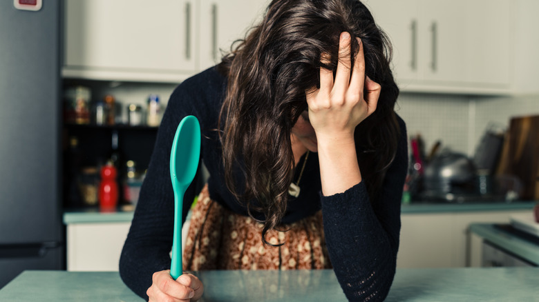 Woman in a kitchen holding her head in frustration, and a spatula