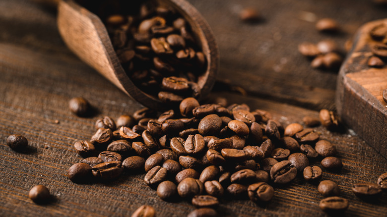 Whole coffee beans in wooden scoop spilling out onto wood surface.