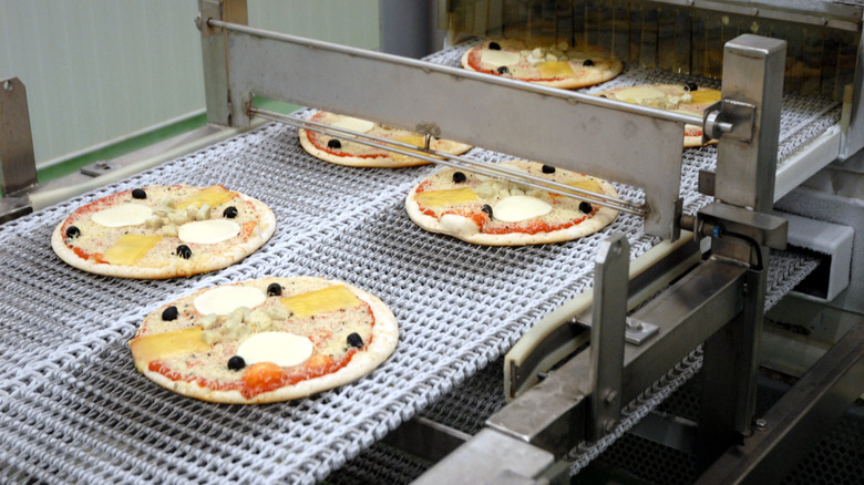 Uncooked pizzas on a conveyor belt going through metal machinery in a factory