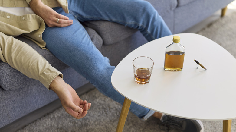Person lying on blue couch atop gray carpet next to white table with brown liquor in bottle and glass