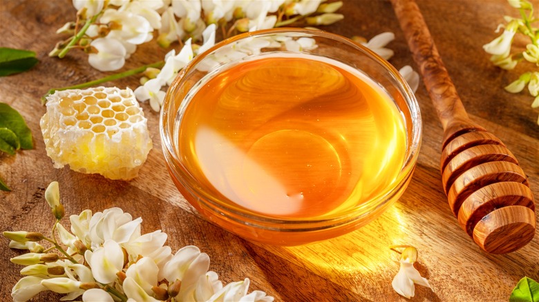A small glass bowl of honey on a wooden board, displayed between a honeycomb and a dipper.