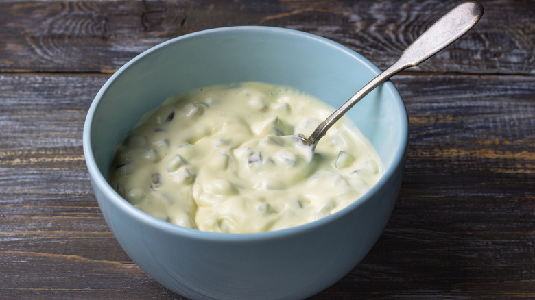 A blue bowl of tartar sauce with a teaspoon inside, on top of a dark, wooden surface