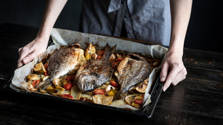 A cook's hands carrying a baking dish of several baked whole fish and vegetables