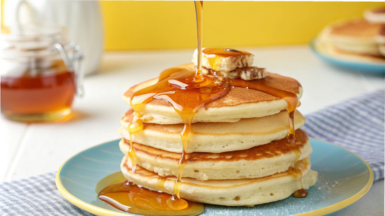 A stack of pancakes topped with pats of butter, maple syrup being drizzled overtop