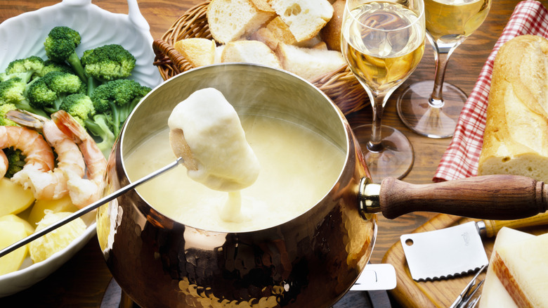 Bread being dipped into a fondue pot surrounded by vegetables, seafood, and wine