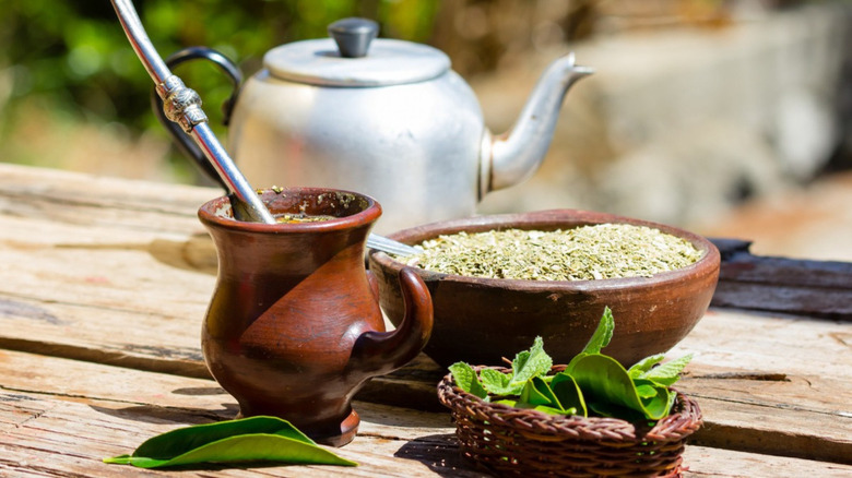 a bowl of yerba mate leaves next to a cup of mate with a metal straw filter, and a teapot in the background all placed on a wooden table. 