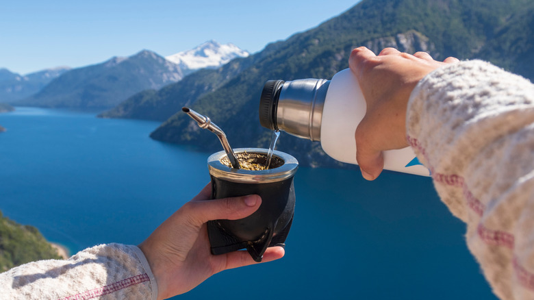 a hiker pouring warm water from a thermos into a cup of yerba mate with a straw. background has scenery of mountains and a clear lake