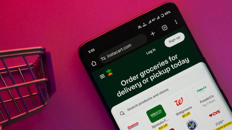 Closeup of a smartphone displaying Instacart.com showing various retailer logos on a pink background