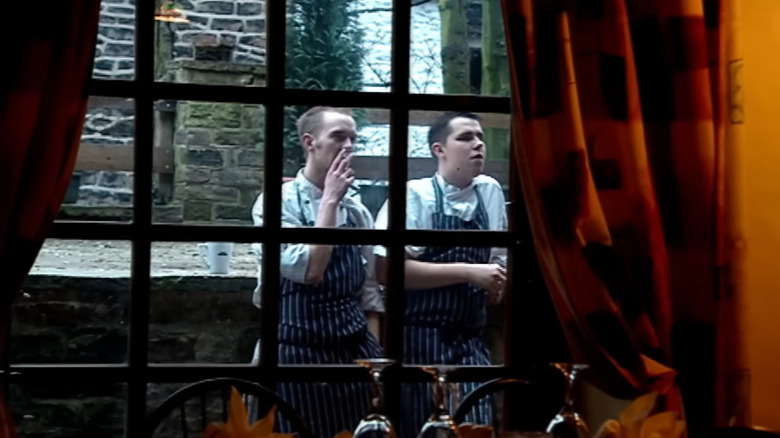 Two cooks stand smoking outside a restaurant window with a stone wall behind them