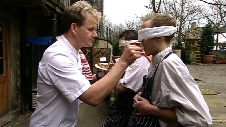 Gordon Ramsay hand-feeds a piece of steak to a cook who is blindfolded on a sidewalk