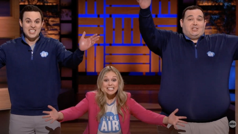 Two men in blue shirts raising their hands up with a woman in a pink shirt in between kneeling slightly and raising her hands to her sides