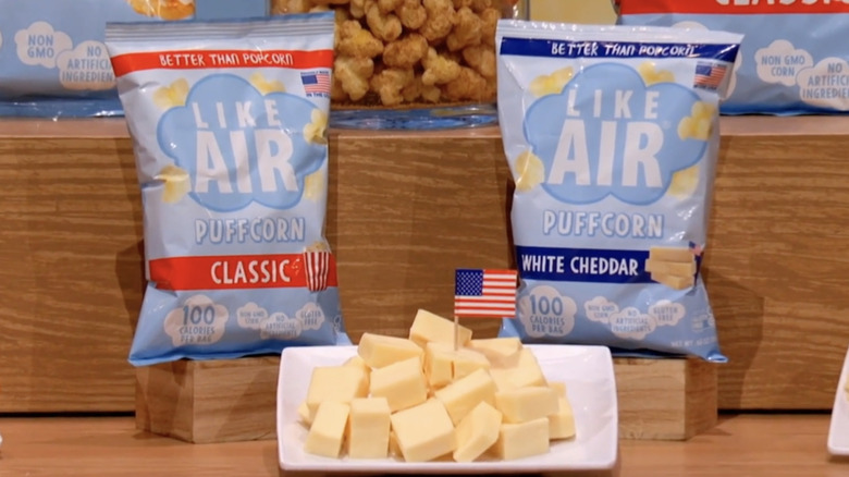 A bag of Like Air Puffcorn Classic and a bag of Like Air Puffcorn White Cheddar with a plate of cheese cubes in between them.